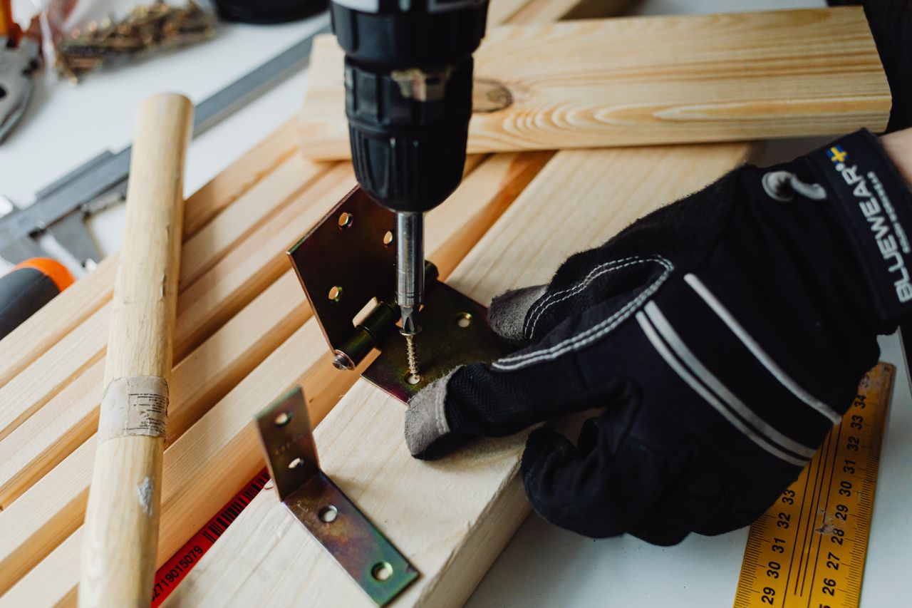 Person wearing work gloves using an electric drill to drive a screw into a metal bracket on a wooden board